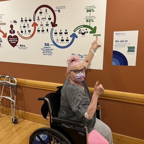 Katrina sits in a wheelchair wearing a mask as she continues her recovery from a heart transplant. She has wavy hair dyed light pink, light skin, and black glasses. From her wheelchair, she reaches up to point at a big diagram of the rehabilitation process on the wall that ends in the word Home.