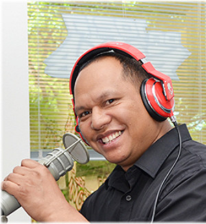 Client Eric Bunda aka DJ KoolE is pictured with headphones and a professional microphone. He has light brown skin, short black hair, and dark eyes and a big smile.