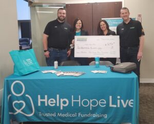 Shannon and Kelly from Help Hope Live hold up a giant check from Express 4x4 Truck Rental for $5,280 made out to Help Hope Live. Two Express staff members stand beside Kelly and Shannon wearing black polo shirts. In front of them is a Help Hope Live booth table with a teal tablecloth, brochures, and pens.