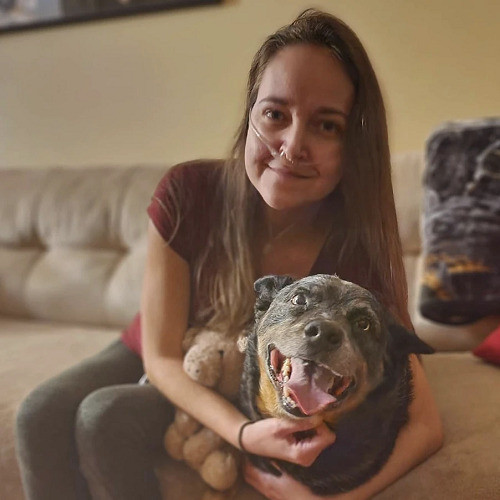 Lung transplant recipient Malwina sits on a couch and hugs an Australian shepherd dog and a teddie bear. She has light skin, long brown hair, and an oxygen tube connected to her nose in this pre-transplant photo.