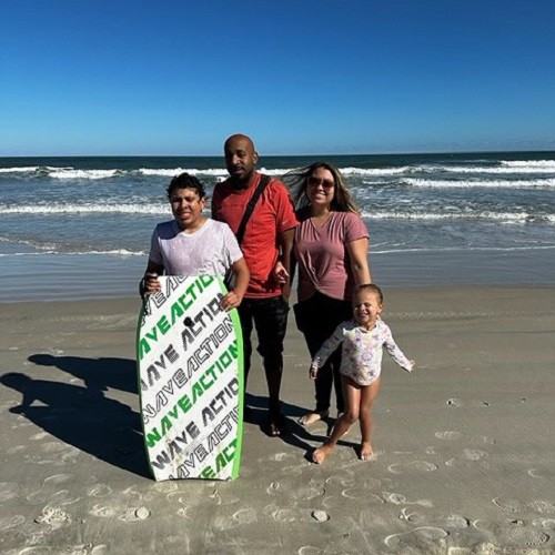 Heart transplant recipient Marcus is pictured with his family on the beach. He has brown skin, a bald head, and a red t-shirt.