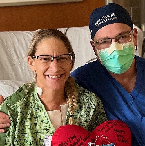 Heart and lung recipient Shawna is in a hospital bed next to a surgeon wearing his mask and scrubs. She holds a heart-shaped signed plushie and has light skin, blonde hair in a fishtail braid, glasses, and a green hospital gown.