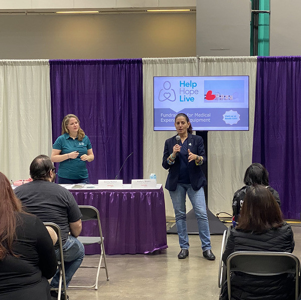 Kelly L Green holds the microphone as she presents about Help Hope Live's medical fundraising to a crowd of people who are either seated in chairs or in their wheelchairs at Abilities Expo LA. Sonny Mullen wears a teal Help Hope Live polo shirt and prepares to add to Kelly's remarks.