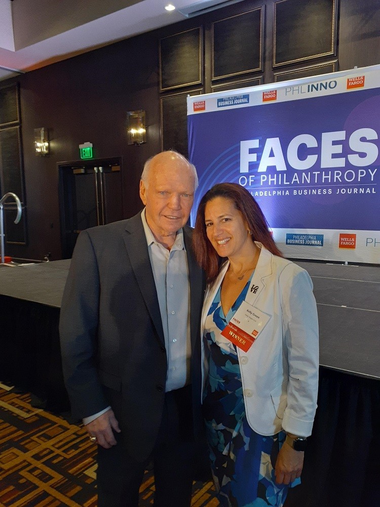 Philadelphia Flyers Alumni Bob Kelly with Help Hope Live Executive Director Kelly L Green at the Philadelphia Business Journey Faces of Philanthropy event. Bob has light skin, blue eyes, and a suit. Kelly has light skin, brown eyes, red-brown hair, and a blue flower print formal dress with a white blazer.