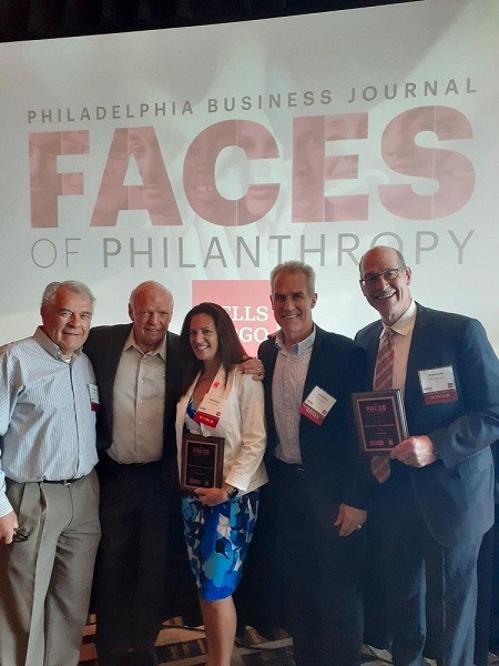 Flyers Alumni Association members Joe Watson, Bob Kelly, Jim Watson, and Brad Marsh stand with Help Hope Live Executive Director Kelly L Green with a large backdrop that reads Philadelphia Business Journal Faces of Philanthropy. The four men wear formal attire and have light skin and gray hair. Holding her award plaque, Kelly has light skin, brown eyes, red-brown hair, and a blue flower print formal dress with a white blazer.