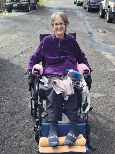Seated in a manual wheelchair, Judy Grossman has gray short hair, light skin, and glasses. She is in the center of a quiet neighborhood street.