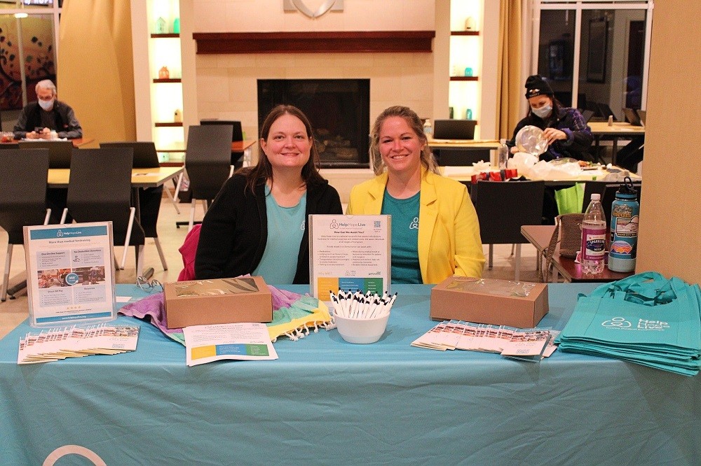 Linda Jara and Help Hope Live's Sonny Mullen are seated at a teal Help Hope Live table at Gift of Life Howie's House. The table features tote bags, informational brochures, pens, and bakery treats. Linda has light skin and brown hair and wears a Help Hope Live teal t-shirt. Sonny has light skin and blonde hair and wears a Help Hope Live t-shirt under her banana-yellow blazer.