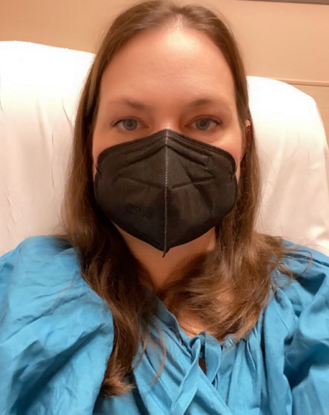In a post-transplant follow-up appointment, Linda Jara wears a blue hospital gown and a black face mask. She has light skin, brown hair past her shoulders, and blue-gray eyes.