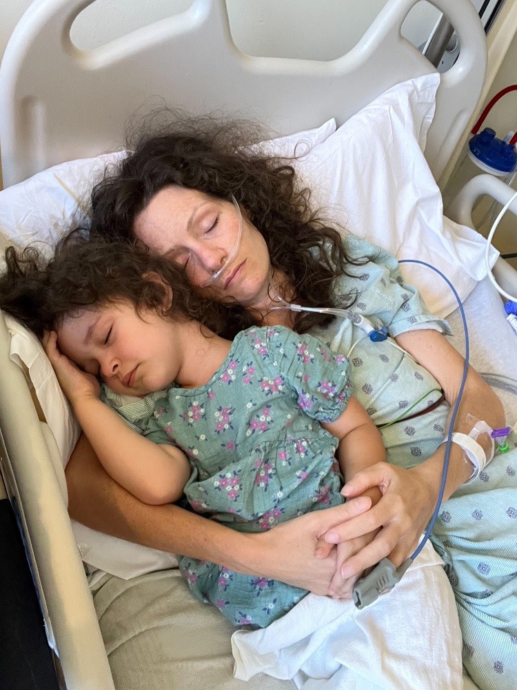 Pamela and her youngest daughter are both asleep in Pamela's hospital bed.. Both have light skin and curly dark brown hair. Pamela wears a hospital gown and has an oxygen tube connected to her nose.