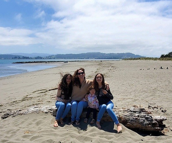 Pamela and her daughters are seated side by side on a log at the beach. They all wear jeans and have light skin and dark hair - some straight and some curly.