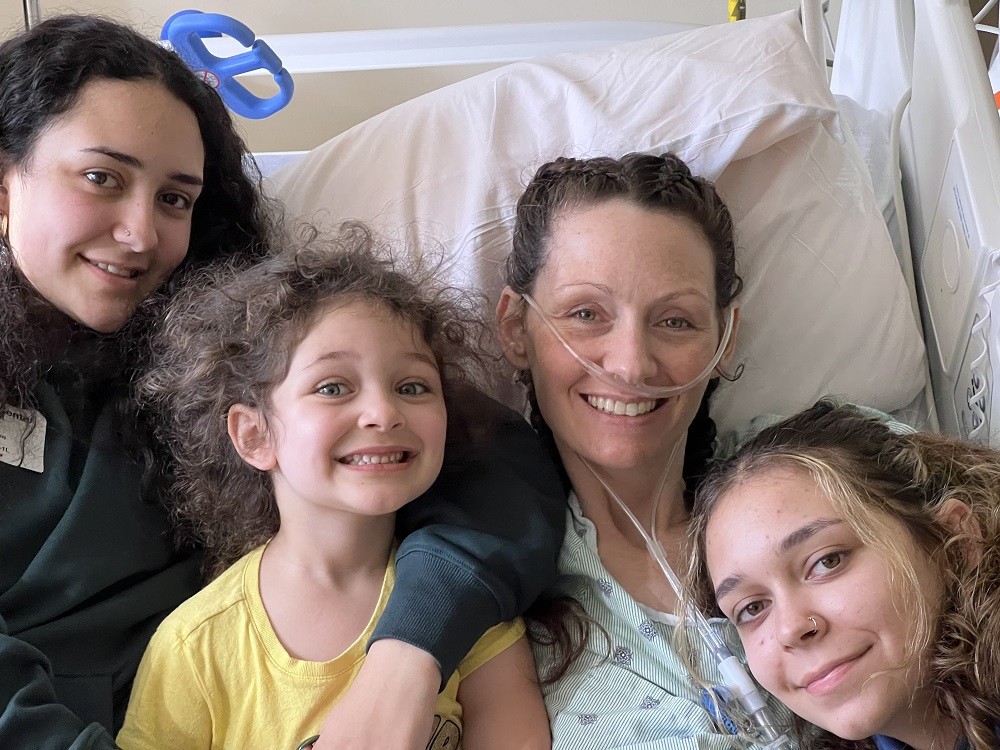 Pamela has light skin and curly brown hair pulled back with an oxygen tube connected to her nose as she sits in a hospital bed with her three daughters. Varying in age, they have light skin and curly brown hair.