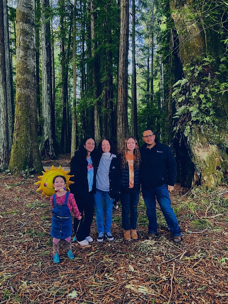 Pamela and her family: two daughters close to her height and close in age, and one youngest daughter, plus her husband. They are wearing outdoor gear in a beautiful forest setting for this informal family portrait.