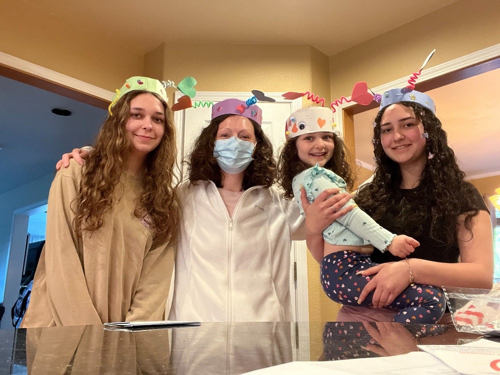 Pamela and her three daughters wear home-made birthday construction paper hats. The first daughter has curly light brown hair. Pamela has a blue face mask and shoulder-length dark brown hair. Her youngest daughter is being held on the hip of an older daughter - both have curly brown hair that looks just like Pamela's.