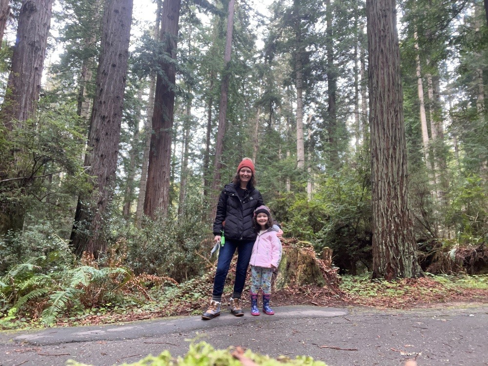 Pamela and her youngest daughter stand on a road with a majestic forest behind them. They are in hiking gear.