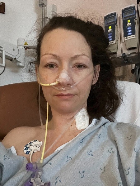 Looking concerned with only a slight smile, Pamela is in the hospital with multiple tubes connected to her body and nose wearing a hospital gown with one shoulder partially exposed. She has light skin, resigned-looking gray eyes, and curly dark brown hair pulled back into a ponytail.