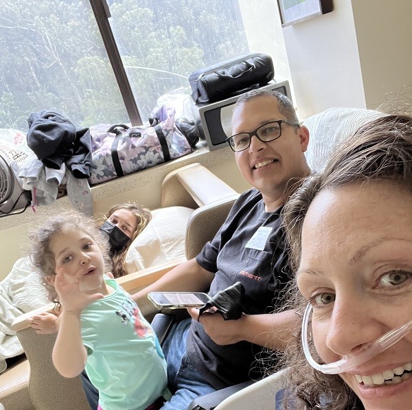 Pamela, two of her daughters, and her husband are together in her small hospital room. She wears an oxygen tube and has light skin and brown hair with gray eyes.