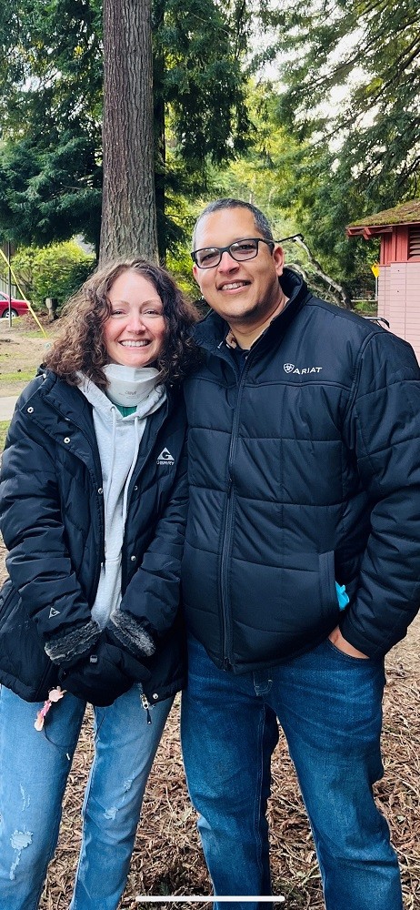 Pamela and her husband wear matching blue jeans and navy-colored quilted outdoor jackets as they stand together outdoors. Pamela has light skin and curly brown hair. Her husband is slightly taller than her with darker skin, glasses, and short-cropped gray hair.