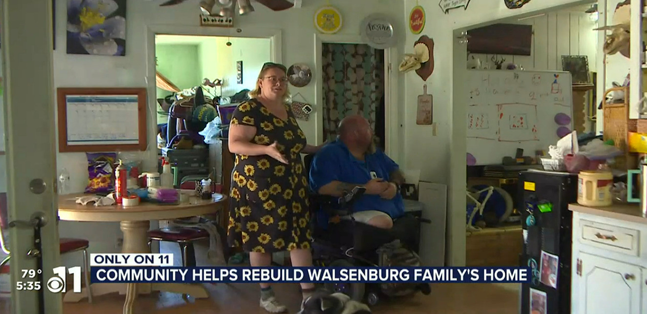 Double amputee Michael Bechaver in his power chair with his wife standing beside him in a floral print sundress in the middle of their home.