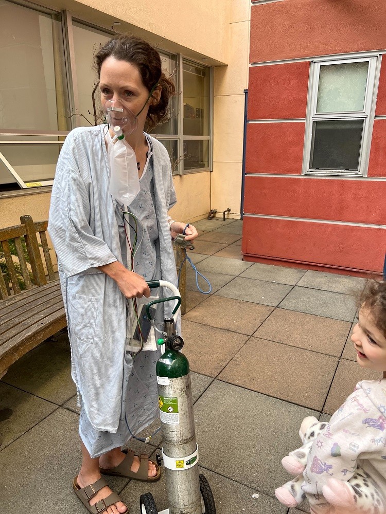 Pamela is in a small courtyard and wears a hospital gown and sandals as she wheels an oxygen tank with her. An oxygen mask connected to the tank covers her nose. She has light skin and curly brown hair pulled back and her youngest daughter is visible at the end of the photo.