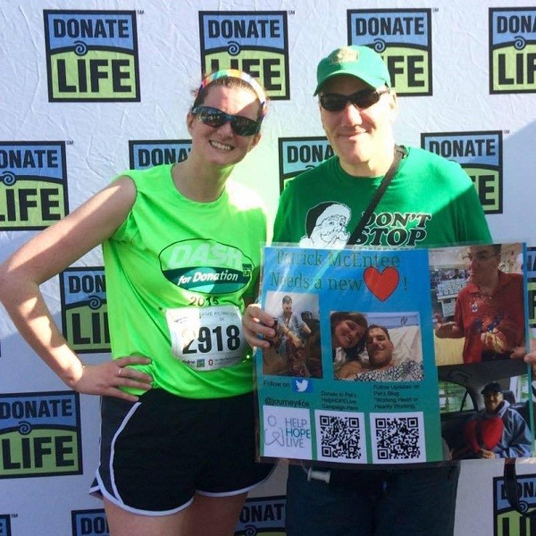 Pat McEntee is pictured pre-transplant attending an athletic event with wife Amy. Behind them is a Donate Life step-and-repeat. Pat and Amy both have light skin, green shirts, and sunglasses. Amy wears a marathon/5K event number and shorts. The strap of Pat's LVAD device is visible. He holds a sign with photos of him and Amy, the Help Hope Live logo, and QR codes.