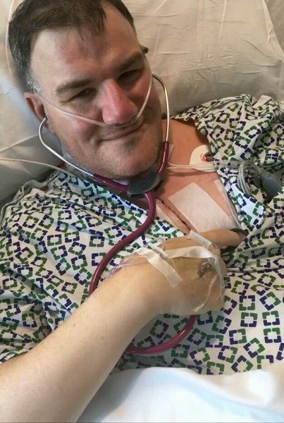 Pat has light skin, gray hair, and a breathing tube connected to his nose as he lies in a hospital bed with a hospital gown. He is listening to his new donor heart beating with a stethoscope and smiling.