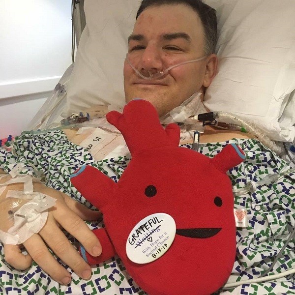 Pat lies in a hospital bed and holds a red anatomical heart plushie with a sticker that has the word WAITING crossed out and replaced with the word GRATEFUL. Pat has light skin, an oxygen tube connected to his nose, and a hospital gown.