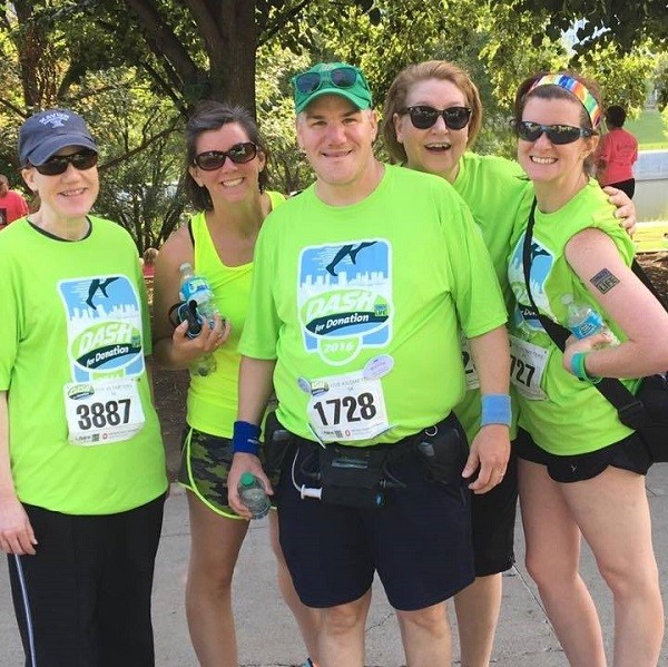 Pat McEntee is pictured pre-tranplant with his LVAD device, wife Amy, and three other women. All are dressed in matching vivid green team t-shirts and wear race numbers for a 5K or marathon.