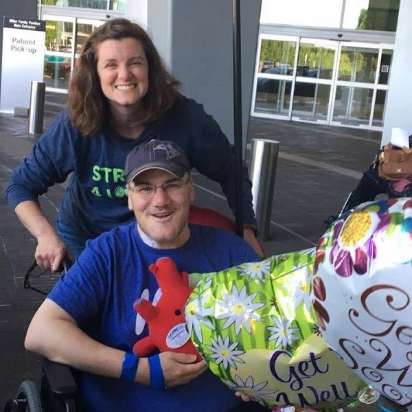 Post-heart transplant, Pat McEntee sits in a wheelchair holding a red heart plushie. He has light skin, glasses, and a blue t-shirt. He is being pushed by his wife Amy, who has light skin and brown hair to her shoulders. Before them are two Get Well balloons.