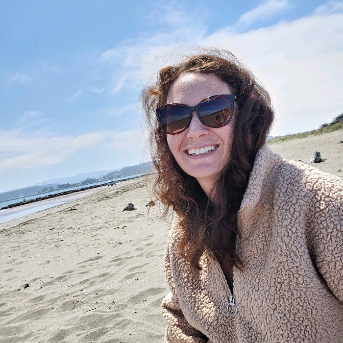 In a selfie at the beach, Pamela has light skin, curly brown hair past her shoulders, brown sunglasses, and a big smile.