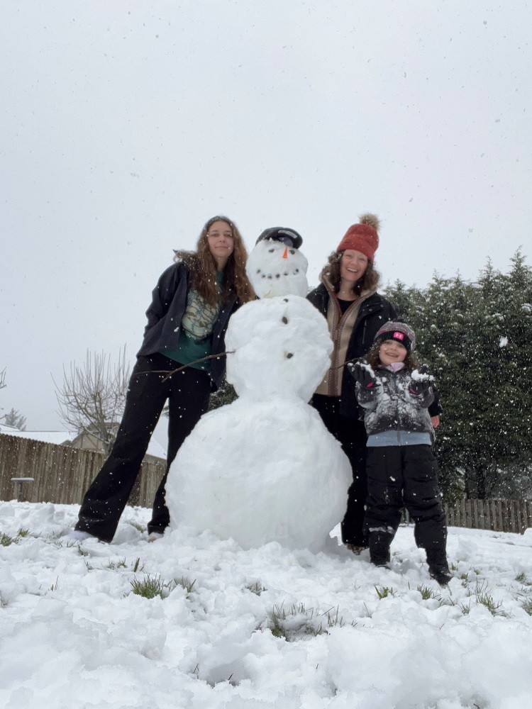 Pamela and two of her daughters are in winter gear and pose next to the snowman they have created, which is as tall as Pamela. All three have light skin and brown hair.