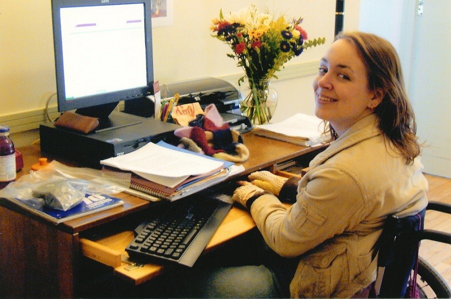 Cindy's daughter Beth is seated in her manual wheelchair at a desk with a computer, keyboard, printer, and paperwork. Beth has light skin, gold hoop earrings, a khaki jacket, and brown wavy hair to her shoulders.