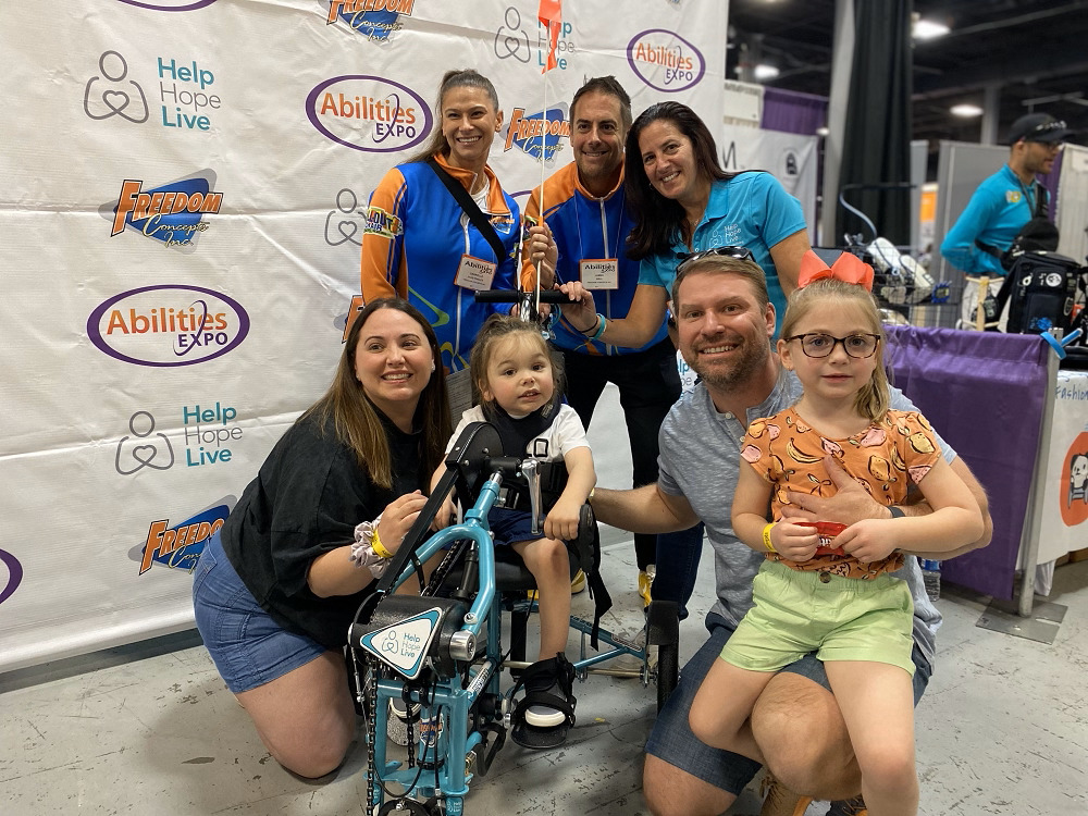 Help Hope Live’s Kelly Green with the family of 3-year-old Easton Clark and Freedom Concepts staff members at Abilities Expo NY. Easton is seated and strapped into a teal Help Hope Live-themed adaptive tricycle.