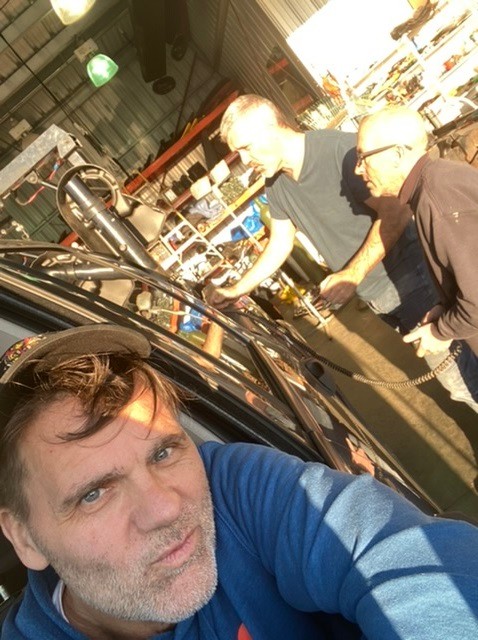 In a selfie, Frank is seated in his vehicle as two men in a shop setting discuss repairs.