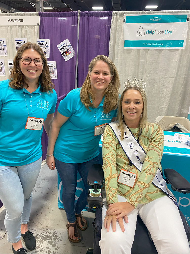 Kate and Sonny are in front of the Help Hope Live booth of Abilities Expo NY Metro with client and Ms Wheelchair America Ali Ingersoll. Kate has light skin, curly brown hair, and glasses and wears a Help Hope Live teal polo. Sonny has sandy hair just past her shoulders, light skin, and a matching polo. Ali Ingersoll has light skin, blonde hair just past her shoulders, and a sparkling tiara with a Ms Wheelchair America sash across her chest. She is seated in a black power chair.