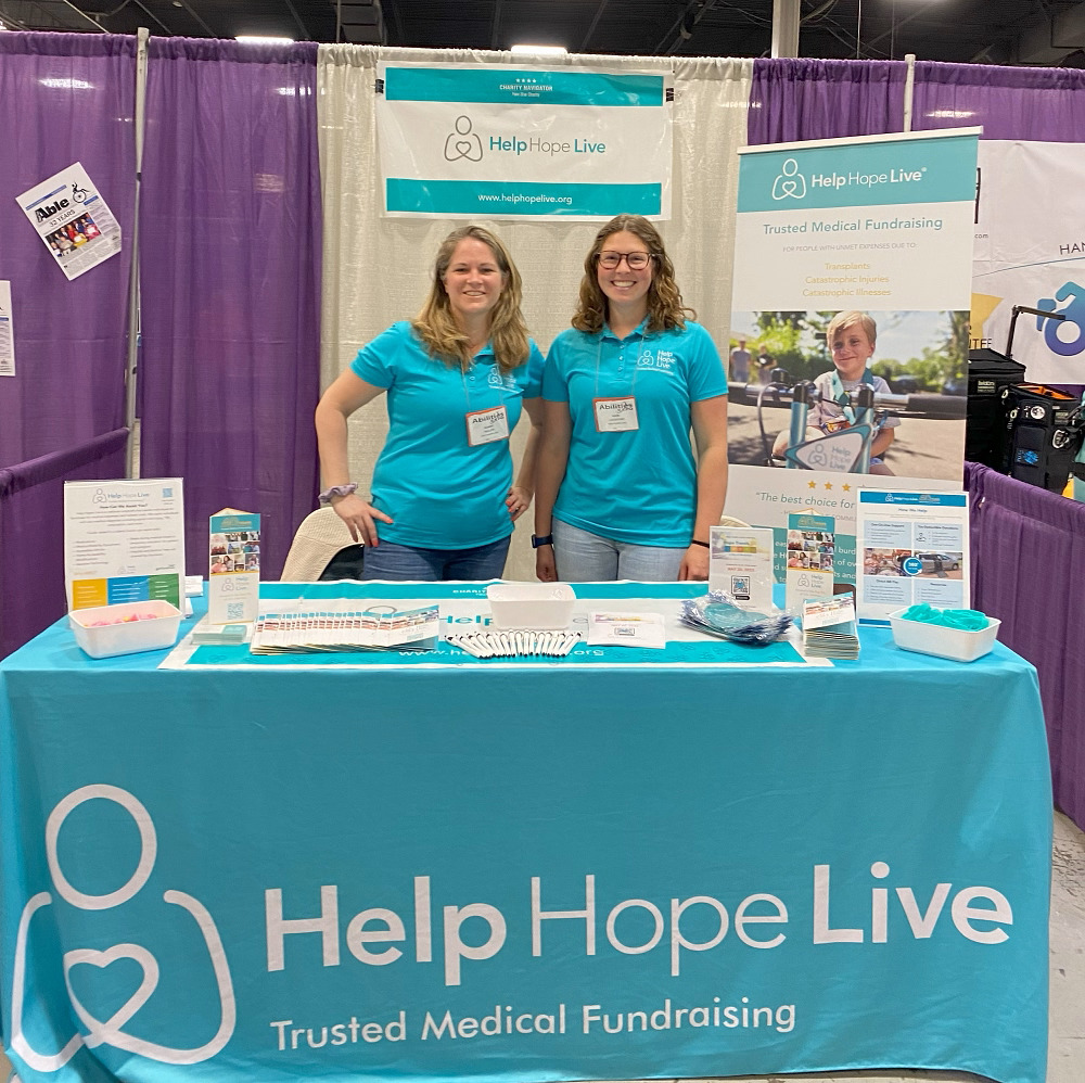 Sonny and Kate from Help Hope Live are at the Help Hope Live booth at Abilities Expo New York Metro. The booth includes a teal tablecloth that reads Help Hope Live: Trusted Medical Fundraising, standing signs and banners, brochures, pens, wristbands, and more. Sonny has light skin and sandy blonde hair. Kate has light skin, glasses, and light brown wavy hair. Both women wear Help Hope Live polo shirts.