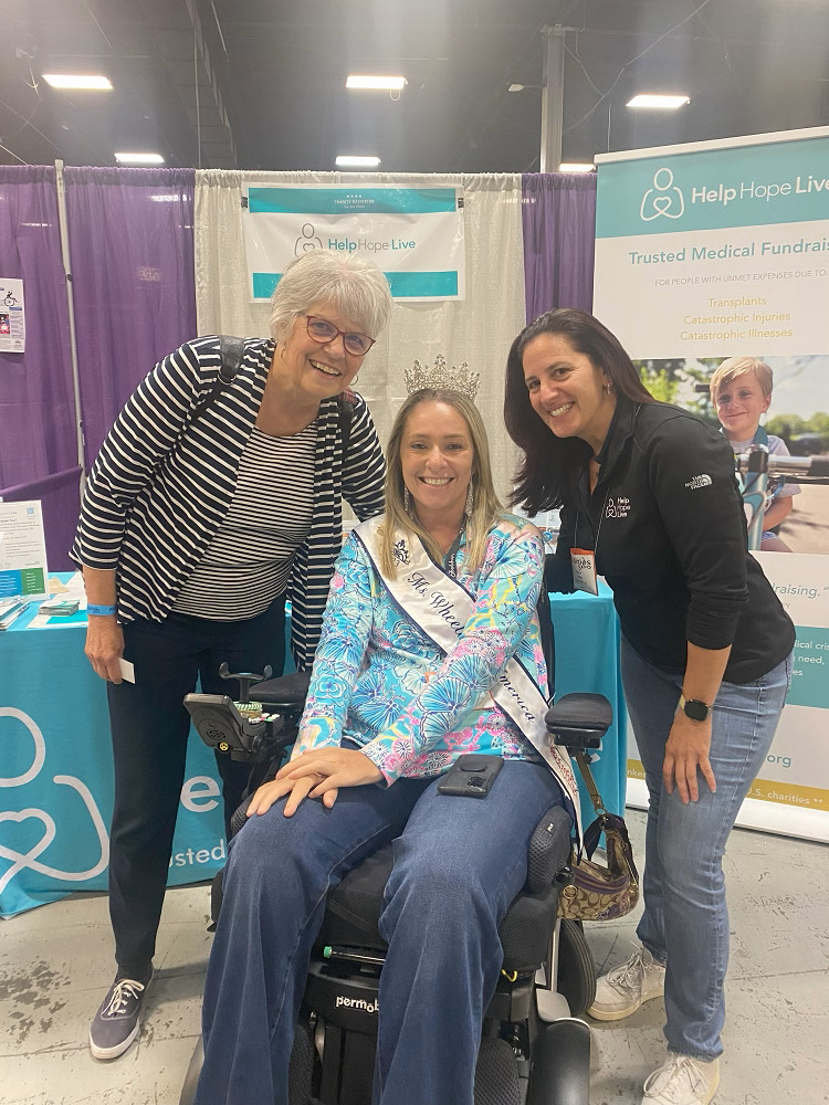 Kelly Green and a Help Hope Live supporter pose in front of the Help Hope Live booth of Abilities Expo NY Metro with client and Ms Wheelchair America Ali Ingersoll. The supporter is a woman with short gray hair, light skin, and glasses. Kelly Green has dark hair past her shoulders, light skin, and a black Help Hope Live North Face jacket. Ali has a vivid printed shirt with a Ms Wheelchair America sash over it as she sits in her black power chair. She has light skin, blonde hair, and a tiara.