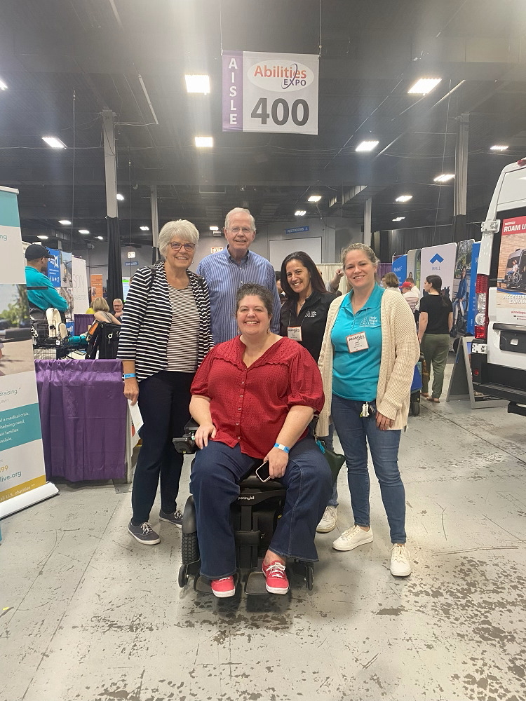 At Abilities Expo NY Metro, Sonny and Kelly pose with Help Hope Live supporters the Murray family. A man and a woman are standing with light skin and gray hair and one woman is seated in her black power chair with light skin, a red blouse, and curly brown hair.