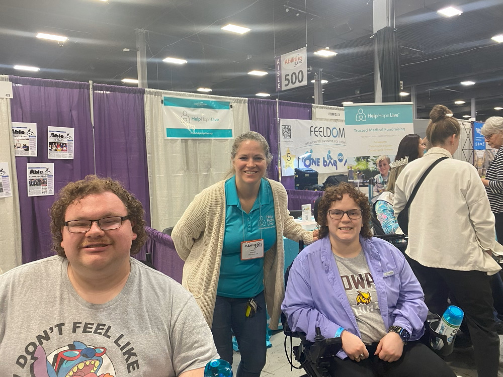 Sonny Mullen is at Abilities Expo NY Metro with Help Hope Live clients Nathan and Charlotte Borgersen. Sonny has light skin an a teal Help Hope Live polo. Nathan has light skin, glasses, and short curly red hair. Charlotte has light skin, curly brown hair, and glasses. The siblings are both seated in their power chairs.