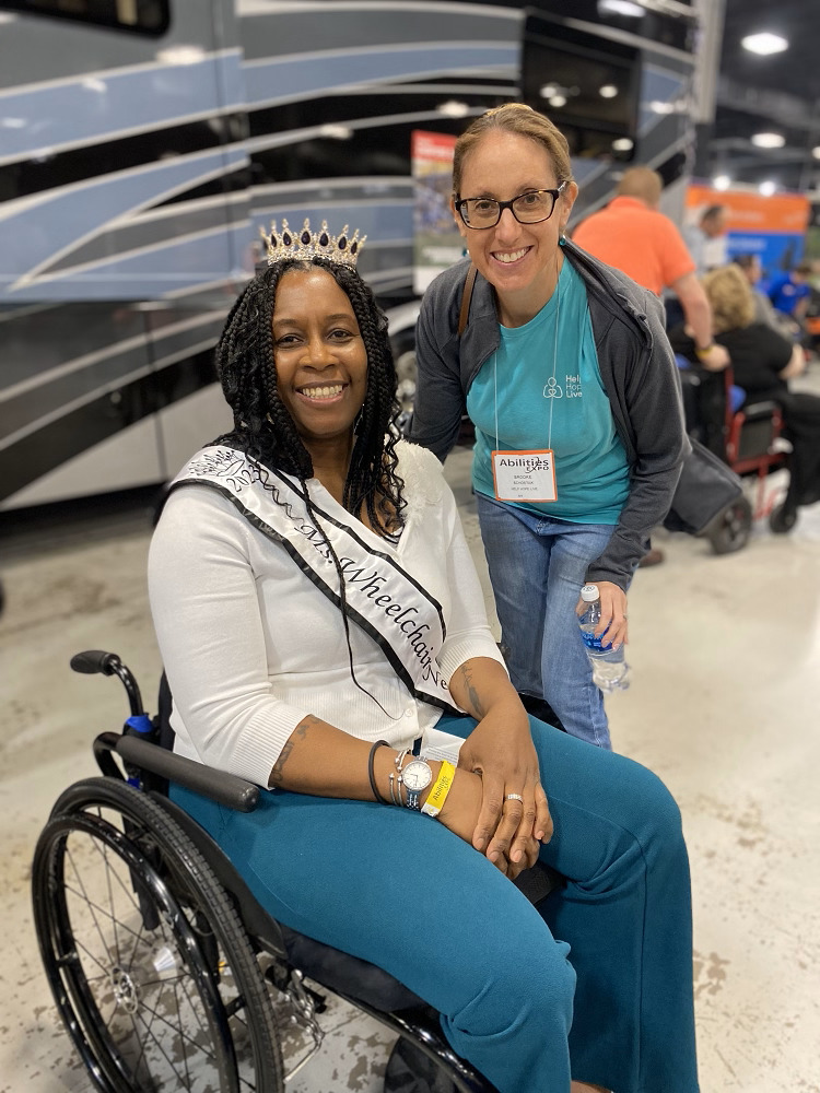 Help Hope Live’s Brooke Schostak is with client Julie Chambers at Abilities Expo NY Metro. Brooke has light skin, blonde hair, dark eyes, and glasses and wears a teal Help Hope Live t-shirt and Abilities Expo nametag. Julie has brown skin, braided black hair, and a sparkling tiara and sash that partially reads Ms. Wheelchair as she is seated in her black manual wheelchair.