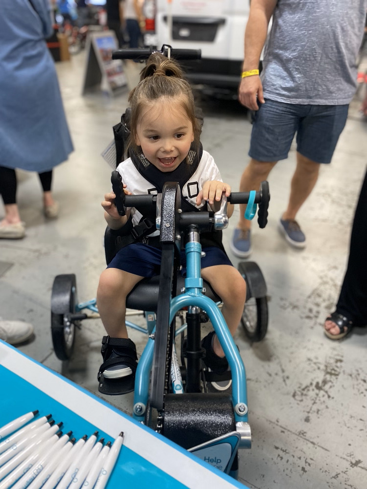 3-year-old Easton sits on his teal adaptive bike at Abilities Expo NY Metro. He has light skin, brown eyes, and long light brown hair.