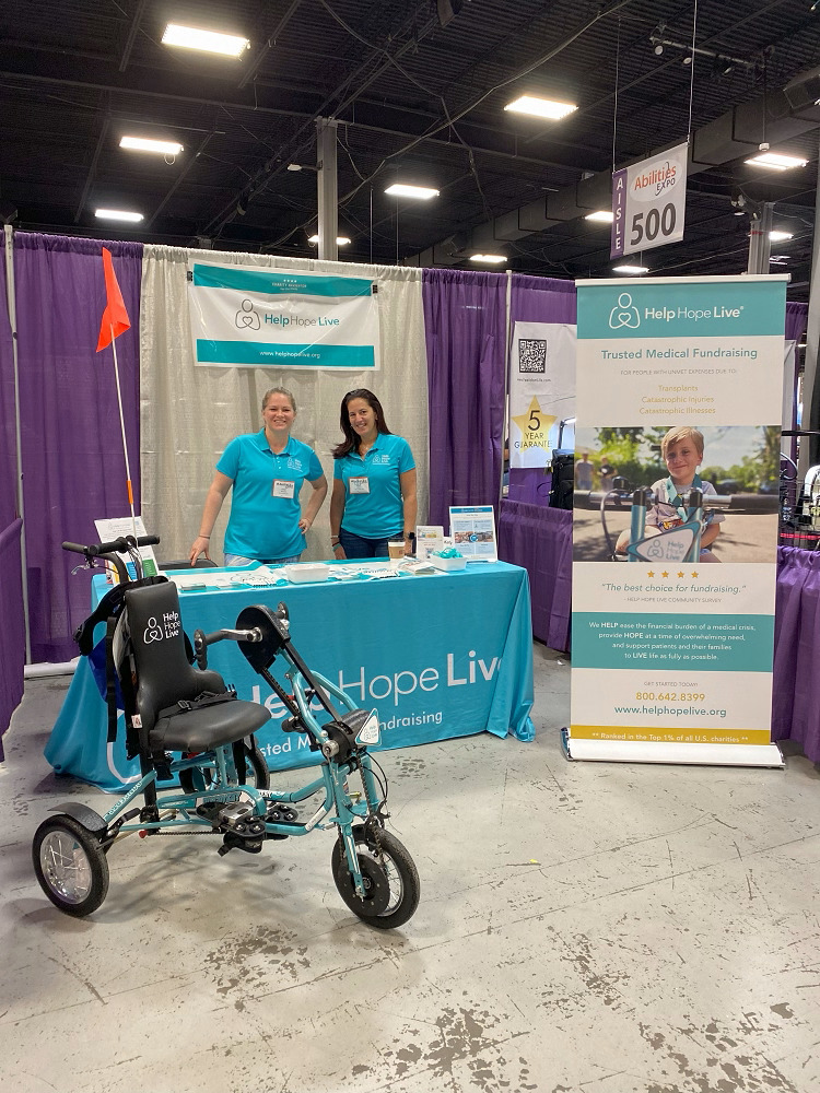 At the Help Hope Live booth of Abilities Expo NY Metro, Sonny Mullen and Kelly Green wear matching Help Hope Live teal polos behind brochures, wristbands, and printed materials. In front of the booth is the Freedom Concepts Help Hope Live-themed teal adaptive bike. Help Hope Live’s logo is stitched onto the backrest in white.