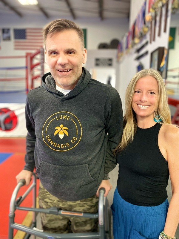 Spinal cord injury survivor Frank Guirlinger is pictured using a walker upright. He has light skin and gray hair. The woman next to him wears athletic gear and has light skin and blonde hair.