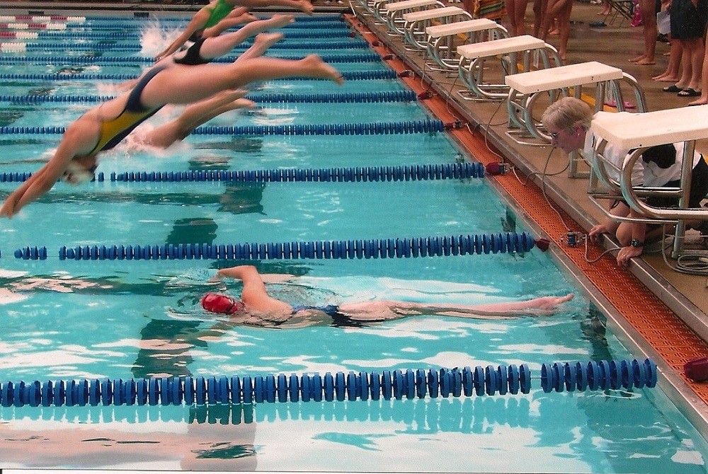 An indoor Olympic-style pool with six lanes of swimmers diving rapidly into the water while one swimmer with a red cap pushes off from the wall in the water and a coach watches.