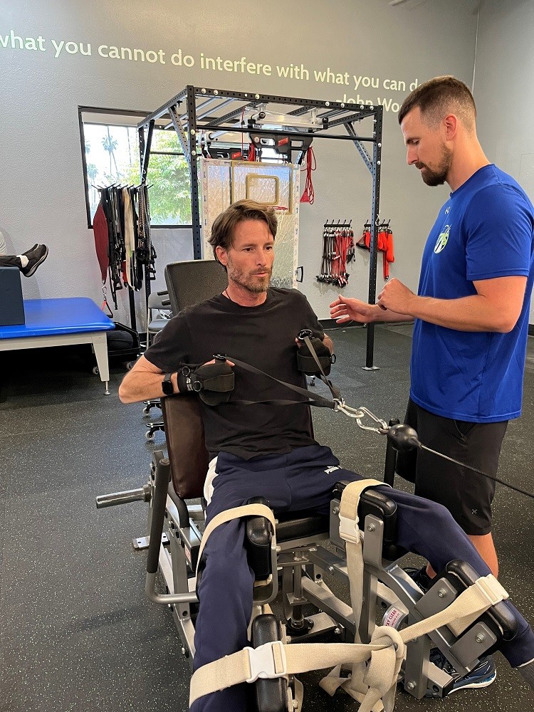 In a rehab or physical therapy environment, Arthur receives assistance from a standing trainer in a bright blue t-shirt as he uses a piece of exercise equipment with his arms. Arthur has light skin, short brown hair, and a short beard and wears comfortable athletic clothing.