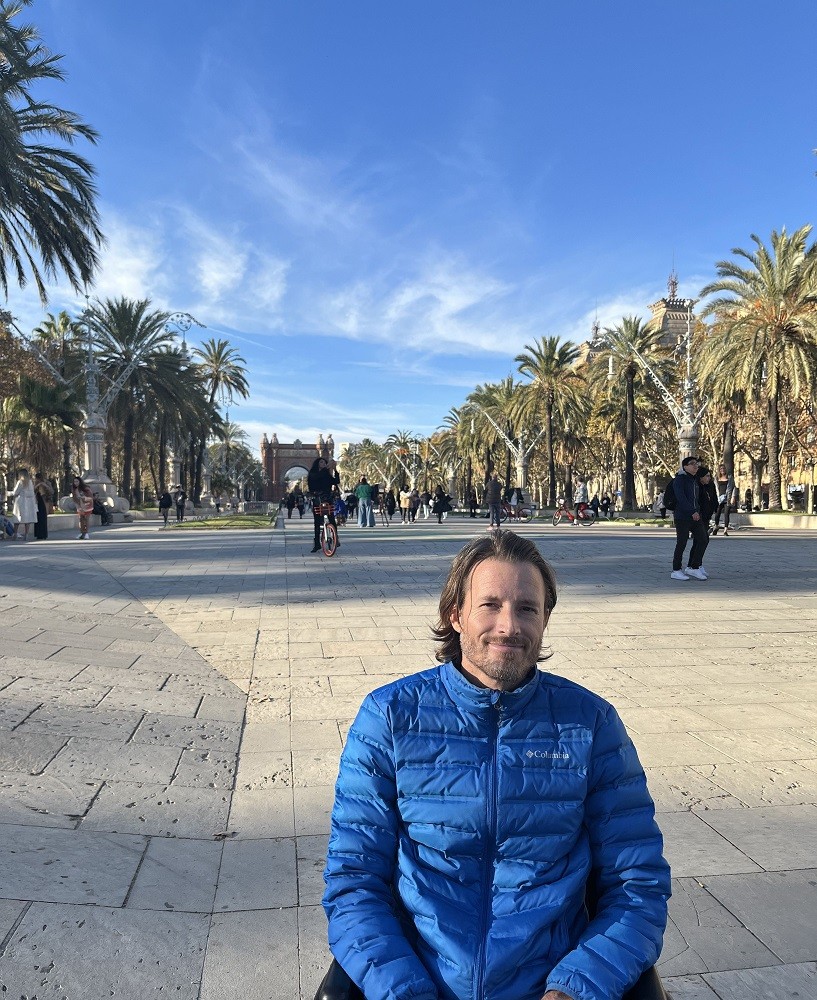 Arthur is seated in his wheelchair in a beautiful outdoor promenade with plenty of walking pedestrians and bikers occupying the tiled roadway lined with gorgeous palm trees. Arthur wears a blue quilted Columbia jacket and has light skin, brown hair to his shoulders, and a short gray and brown beard.