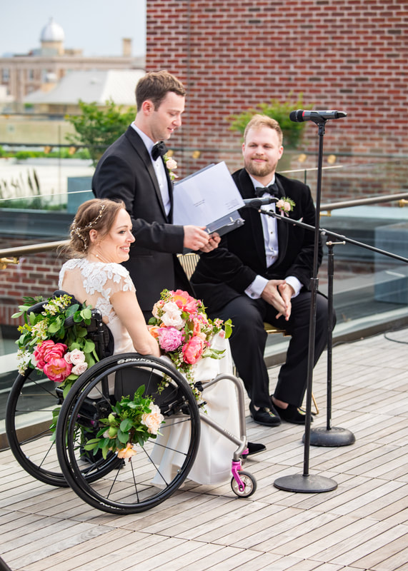 Mid-ceremony, Beth smiles at the crowd during a rooftop marriage ceremony. She is seated in a black manual wheelchair with gorgeous florals attached to the back and wheels that match the blooming bouquet in her lap. The back of her white wedding dress is visible with lace cutouts. Her groom is seated in black formal attire and a velvet jacket. An officiant stands between them in black formal attire.