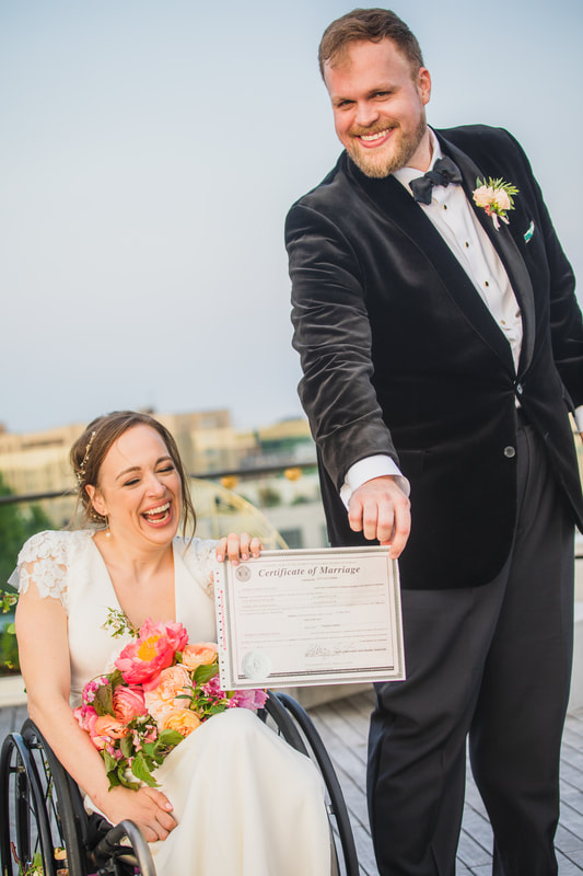 Beth holds a marriage certificate with her husband as they both grin on a rooftop. Beth is wearing her white bridal gown and holding a gorgeous bouquet of multi-colored blooms on her lap as she sits in a black manual wheelchair. Her groom is standig with a black velvet suit top and formal attire.