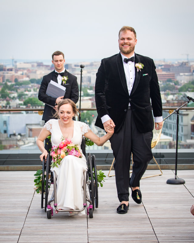 Beth holds her groom's hand as they walk and roll away as a married couple from the officiant. They are on a rooftop. Beth's groom has light skin, light brown hair and a beard, and a black velvet suit top with tassled loafers. Beth wears a white wedding gown with a V-neck and holds a floral bouquet on her lap. She is seated in her black manual wheelchair.