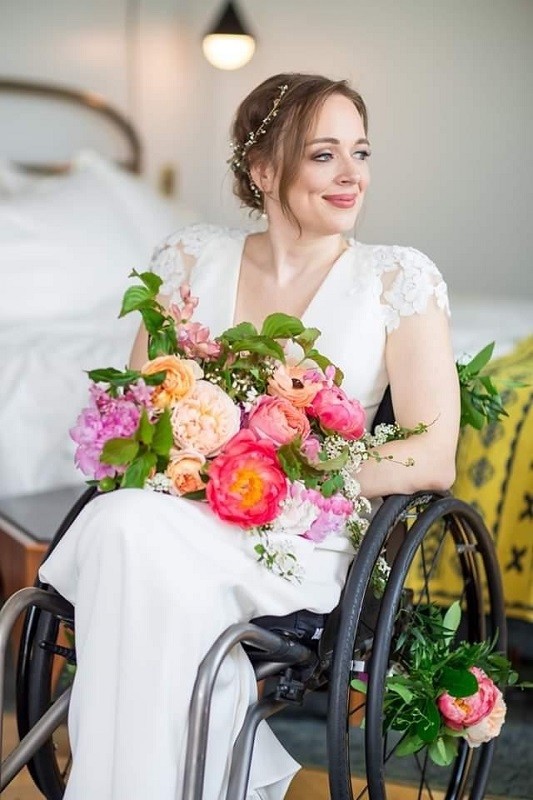 Cindy's daughter Beth is stunning in a wedding portrait. Her light brown hair is in a formal style with a jeweled vine-like accessory. Beth has formal makeup and light skin and wears a white wedding dress with lace short sleeves. She holds a beautiful bouquet of peonies and other flowers in pink shades. There is a matching mini-bouquet attached to the center of the wheels of her manual wheelchair.