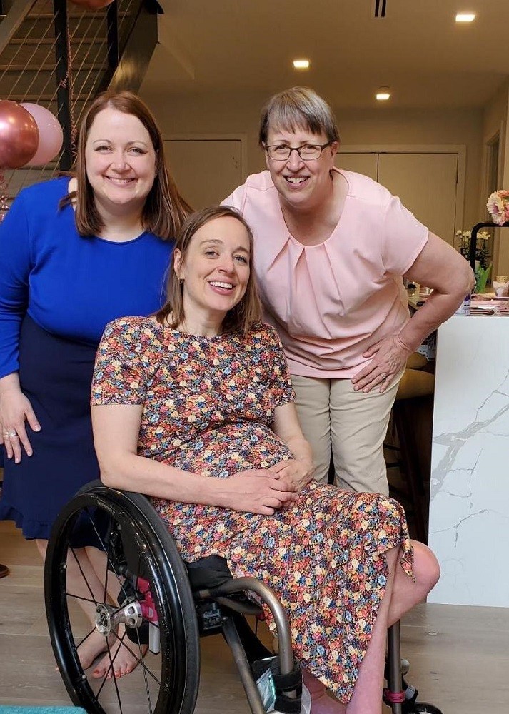 Cindy Kolbe leans down for photo with her daughter, Beth, at a baby shower. Cindy has light skin, glasses, short gray hair, and a pink blouse. Next to her, a smiling woman has shoulder-length red hair, light skin, and a blue dress. Cindy daughter Beth is seated in her black manual wheelcahir and wears a flower print dress. She has light skin, shoulder-length brown hair, light eyes, and a growing baby bump.